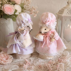 May include: Two fabric mouse figurines dressed in pastel clothing. The left mouse wears a lavender bonnet and dress, holding a small baby mouse. The right mouse wears a pink bonnet and dress, holding a small plate with a round white pastry.
