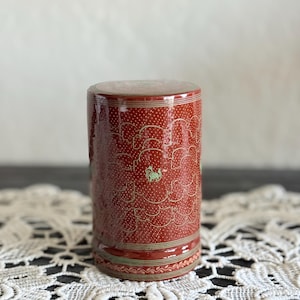 May include: A cylindrical, red ceramic container with a lid. The container is decorated with a repeating pattern of white dots and gold cloud-like shapes. A small green animal figure is visible on the side.