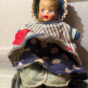 Puede incluir: Muñeca vintage con rostro pintado, ojos azules y labios rojos. La muñeca viste un gorro y un vestido con rayas azules y blancas, una falda de lunares azules y un parche rojo. Una muñeca de colección clásica.