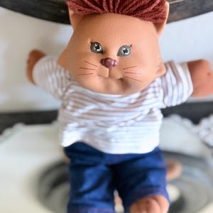 May include: A vintage doll in the shape of a cat with a brown face, red yarn hair, and brown arms and legs. The doll is wearing a white and light brown striped shirt and blue jeans. The doll has painted eyes and a pink nose.