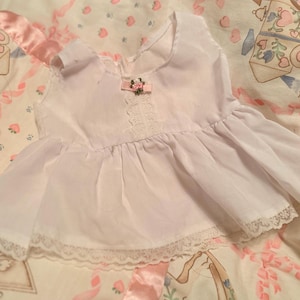 Op de afbeelding: Een witte baby-doll top met een ronde halslijn, korte mouwen en een geplooid zoom. De top heeft een kleine roze strik met een bloemenaccent en kanten details op het lijfje. Het kledingstuk is gemaakt van een lichte stof.