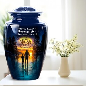 May include: A dark blue cremation urn with a glossy finish, featuring a printed scene of two people fishing in a stream at sunset. The text reads: In Loving Memory of Racheal John. Dec/1956 - Jan/2025. IN THE STREAMS OF MEMORY YOU FISH ON FOREVER.