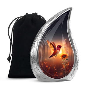 May include: A silver-colored teardrop-shaped urn with a detailed floral design. The front features a vibrant image of a hummingbird in flight near a sunflower against a sunset backdrop. Includes a black drawstring bag.