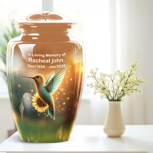 May include: A decorative urn with a hummingbird and sunflower design. The urn is a light brown color with a lid. The text "In Loving Memory of Racheal John. Dec/1956 - Jan/2025" is printed on the front.