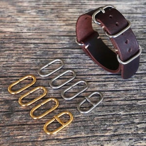 May include: A dark brown leather watch strap with silver-tone hardware is displayed next to several metal watch strap loops. Some loops are gold-toned, and others are silver-toned. The items are arranged on a weathered wooden surface.