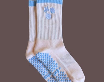 May include: A pair of light pink socks with blue accents. The socks have a ribbed texture and feature blue seashell embroidery near the top. The soles have a blue, heart-shaped, non-slip grip.