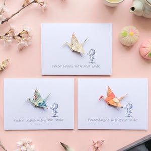 Op de afbeelding: Drie witte wenskaarten met origami-kranen in beige, blauwgroen en oranje. Elke kaart heeft een kleine illustratie van een persoon en de tekst "Peace begins with your smile."