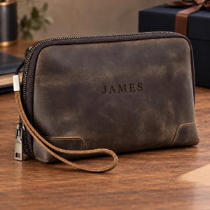 Op de afbeelding: Een donkerbruine leren tas met ritssluiting, polsband en de naam JAMES in het midden gegraveerd. Het rechthoekige accessoire heeft een gladde, matte afwerking met zichtbare stiksels in de onderste hoeken en een metalen ritssluiting.