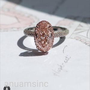 Peut inclure: Une bague en diamant avec une grosse pierre rose de taille ovale. La bague a un halo de petits diamants et un anneau incrusté de minuscules diamants. La bague est posée sur une surface blanche.