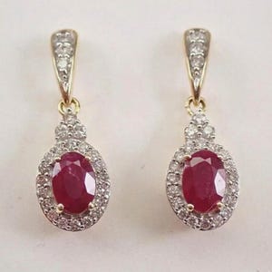 May include: A pair of gold earrings with oval, deep red gemstones, encircled by smaller, clear stones. The earrings have a decorative top section with more clear stones. The earrings are a classic design.