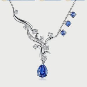 May include: A silver necklace with a floral design. The necklace features clear and blue gemstones. A large, teardrop-shaped blue gemstone hangs from the centre. Smaller blue gemstones dangle from the chain. Clear gemstones are set in the floral design.
