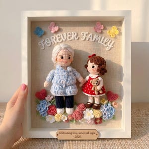 Könnte beinhalten: Ein gerahmtes Häkelkunstwerk mit zwei Puppen, eine in einem blauen Mantel und die andere in einem roten Kleid, die sich an den Händen halten. Die Worte "FOREVER FAMILY" stehen darüber, mit floralen und herzförmigen Akzenten. Eine kleine Holzplakette lautet "Celebrating love, across all ages. - 2026."