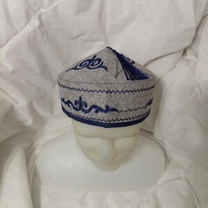 May include: A light gray felt cap featuring dark blue embroidered scrollwork patterns along the sides and top. The hat is finished with a dark blue trim at the base and a dark blue tassel attached to the center of the crown, creating a structured look.