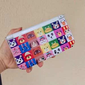 Könnte beinhalten: Eine weiße Handheld-Spielkonsole mit einem farbenfrohen Muster, das verschiedene Cartoon-Tiergesichter zeigt. Das Muster ist ein Raster aus unterschiedlich farbigen Quadraten. Die Konsole hat ein Klappdesign.