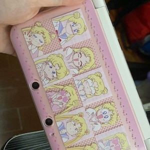 Può includere: Custodia rosa per Nintendo 3DS con diverse illustrazioni del personaggio di Sailor Moon. La custodia ha uno sfondo rosa chiaro con un motivo ripetuto di immagini di Sailor Moon in varie pose. La custodia ha due pulsanti circolari neri e una cerniera bianca.