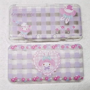 Puede incluir: Dos fundas transparentes de plástico para Nintendo 3DS. Las fundas presentan un patrón de cuadros morados y blancos. Una funda tiene un personaje de dibujos animados con un lazo y las palabras "My Melody". La otra funda tiene un personaje de dibujos animados con un lazo y flores.
