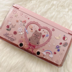 May include: Pink handheld gaming console case decorated with various stickers, including a brown bear inside a heart-shaped frame, flowers, and a rainbow. The word "Happy" is written in cursive on the case.