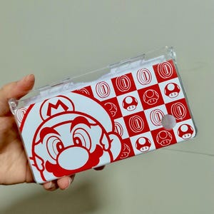 May include: A red and white Nintendo 3DS case featuring a large graphic of Mario's face on the left side. The right side has a checkerboard pattern with red coins and mushrooms. The case is held in a person's hand.