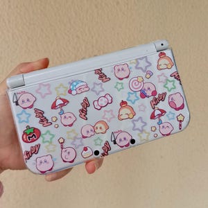 Puede incluir: Una consola de juegos portátil Nintendo 3DS blanca con una cubierta protectora transparente. La cubierta presenta un patrón repetido de personajes rosas de Kirby, estrellas, caramelos y la palabra "Kirby". El diseño incluye el texto "hehe..." en varios lugares.