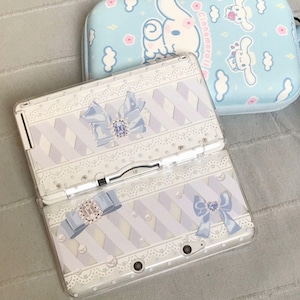 May include: A white and light blue Nintendo 3DS console with a matching case. The console features a decorative design with blue bows, lace, and a striped pattern. The case has a light blue background with cartoon characters.