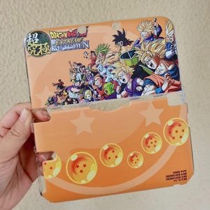 May include: An orange and clear plastic case for a handheld gaming device. The case features a colourful graphic of characters from the Dragon Ball series, with the text "DRAGON BALL EXTREME BUTODEN" and Japanese characters. The design includes stars and dragon balls.