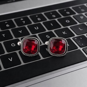 May include: Two square cufflinks featuring a deep red gemstone set within a silver, rope-style frame. The cufflinks are displayed on a dark surface, emphasizing their color and design. A classic accessory.