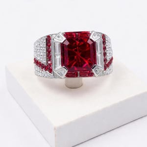 Könnte beinhalten: Ein auffälliger Ring mit einem großen, rechteckigen roten Rubin. Der Silberring ist mit kleineren weißen und roten Edelsteinen in einem geometrischen Muster besetzt. Der Ring wird auf einer weißen Oberfläche präsentiert.