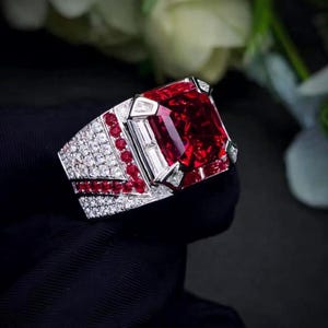 Puede incluir: Un llamativo anillo con una gran gema roja rectangular. La banda plateada está adornada con pequeñas gemas blancas y rojas. La piedra roja central es el punto focal. El anillo está sobre un fondo oscuro.
