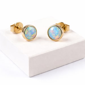 May include: A pair of gold-colored stud earrings with round, iridescent opal stones. The opals display a mix of blue, pink, and white hues. The earrings are displayed on a white surface, showcasing their design and color.