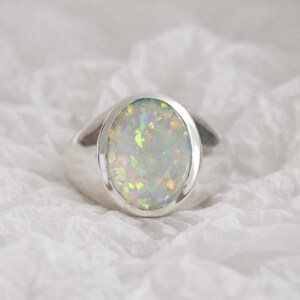 Puede incluir: Anillo de plata con una piedra de ópalo ovalada. El ópalo tiene un color base blanco lechoso con destellos verdes, amarillos y naranjas. El anillo tiene un acabado liso y pulido.