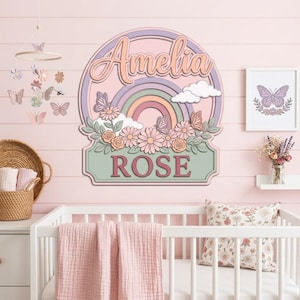 May include: A pastel-coloured nursery wall decor piece with the name "Amelia" above a rainbow and the name "Rose" below. The design includes flowers, butterflies, and clouds. The room also features a cot, mobile, and framed butterfly art.