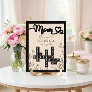 May include: A wooden framed sign with the word "Mom" and a heart design. The sign includes the text "You are the piece that holds us together" and a crossword puzzle with names. The sign is displayed on a wooden stand.