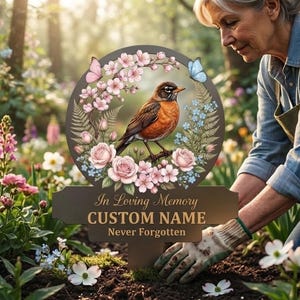 Peut inclure: Un piquet de jardin commémoratif circulaire orné d'un rouge-gorge brun sur une branche, entouré de fleurs de cerisier roses, de roses et de papillons bleus. Le texte indique In Loving Memory, CUSTOM NAME et Never Forgotten en lettres dorées.