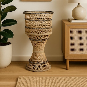 Op de afbeelding: Een geweven plantenstandaard van rotan met een uitlopende voet en een cilindrische bovenkant. De standaard staat in een kamer met een ingelijst abstract kunstwerk, een lamp en een houten kast. De standaard is ongeveer 76 cm hoog.