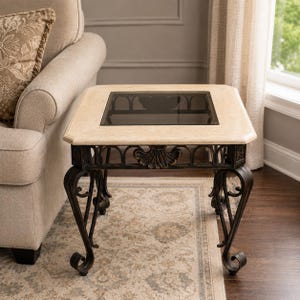 May include: A square side table featuring a beige stone top with a central dark glass inset. The base is constructed from dark metal with ornate scrollwork legs and a decorative shell motif centred on each side of the frame beneath the tabletop.