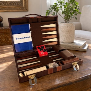 May include: A brown leather backgammon set in an open briefcase style case with a dark brown felt interior. The board features white and brown triangular points, a blue instruction booklet labelled Backgammon, a red deck of cards, and game pieces.