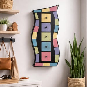 May include: A colorful wall-mounted cabinet with a wavy black frame. It features five square drawers with black knobs, surrounded by pastel-colored mosaic tiles. The cabinet is a decorative storage solution.