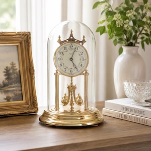 May include: A gold-toned anniversary clock with a circular white face, black numerals, and a rotating pendulum, all enclosed within a clear glass dome. The clock features a decorative crown-like top and sits on a polished gold-coloured base.