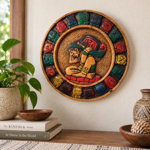 May include: A circular wall plaque featuring a central relief carving of a seated figure in profile, surrounded by a border of colourful, repeating glyph-like symbols in shades of red, green, blue, and yellow against a textured brown background.