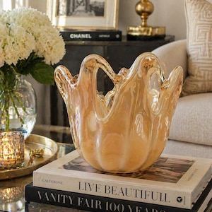 May include: An amber-hued glass bowl with a distinctive, undulating edge. The bowl rests on a stack of books, including "Live Beautifully" and "Vanity Fair 100 Years", on a gold-toned coffee table. The background features a vase of white flowers and a framed picture.