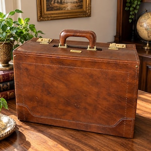 May include: A rectangular brown leather briefcase featuring a sturdy top handle, brass-toned metal latches, and a small rectangular nameplate with the text Stradella centred below the handle. The case has visible stitching along the edges.