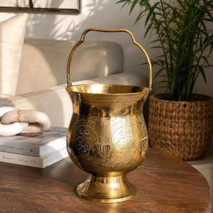 May include: A polished brass bucket with a rounded body, a flared pedestal base, and a curved swing handle. The exterior surface features intricate, etched floral patterns and decorative line work encircling the midsection of the vessel.