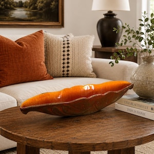 May include: An orange ceramic decorative bowl with a leaf-like shape sits on a round wooden table. The bowl has a glossy finish and a textured, patterned design. The background includes a sofa with pillows and a lamp.