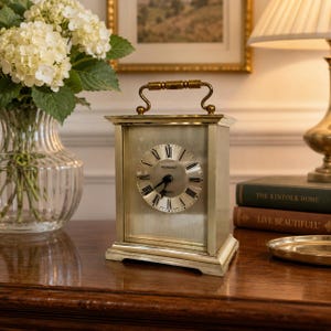 Puede incluir: Un reloj de sobremesa rectangular de latón con un asa superior y una esfera circular con números romanos negros y el texto "PRESIDENT" y "QUARTZ" impreso en el centro. El reloj tiene una esfera plateada metálica y un marco de latón.