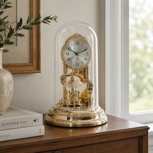 Peut inclure: Une horloge à quartz dorée avec un dôme en verre. Le cadran de l'horloge comporte des chiffres romains et des aiguilles noirs. La base et les éléments décoratifs sont ornés. L'horloge est exposée sur une surface en bois.