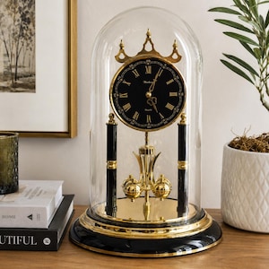 May include: A mantel clock encased in a clear glass dome featuring a black circular face with gold Roman numerals and hands. The clock is supported by two black pillars with gold accents and a rotating gold pendulum base on a black circular stand.