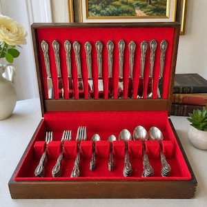 May include: A wooden cutlery storage chest with a vibrant red velvet interior lining. The open case displays a collection of silver-toned metal utensils, including twelve upright knives in the lid and various forks and spoons arranged in the base.