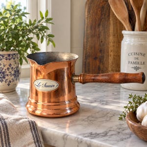 May include: A copper sauce pot with a polished finish, a long wooden handle, and a pouring spout. An oval white label on the front features the text Le Saucier in black script. The interior of the pot is metallic silver, and the base is wide.