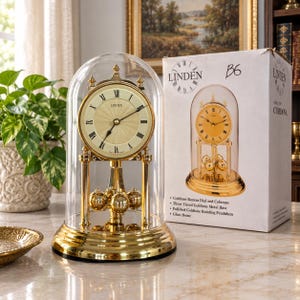 May include: A gold-tone Linden mantel clock featuring a round face with black Roman numerals, a rotating pendulum with four spheres, and a clear glass dome. The clock is displayed next to its white cardboard box labeled Linden B6.