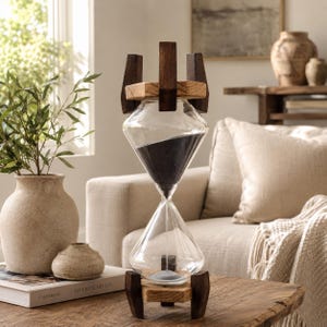 May include: A diamond-shaped glass hourglass featuring dark black sand inside. The hourglass is held within a frame constructed from light-toned wooden platforms and dark brown vertical wooden supports at both the top and the bottom ends.